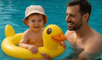 Μικρό αγόρι με καπέλο κάθεται σε σωσίβιο-παπάκι μέσα στην πισίνα, ενώ ο πατέρας του το στηρίζει και του χαμογελά